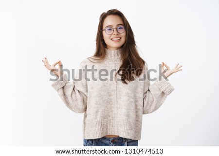 Portrait of happy peaceful good-looking woman in glasses and warm sweater keep calm as meditating smiling broadly at camera raising hands with mudra, zen orbs sharing yoga knowledge over gray wall