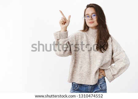 Determined and assertive self-assured good-looking woman in glasses and sweater pointing at upper left corner as making choice and wanting pick thing, standing confident over gray background