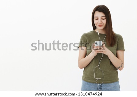 Girl searching album of singer in app wearing wired earphones and typing in smartphone looking casually with pleased happy smile at mobile phone screen, watching video in headphones not disturb others