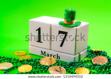 St Patrick's Day meme and Irish spring holiday concept theme with a block calendar showing the date of March 17, leprechaun hat with sparkling clover leaf, shamrock, beads isolated on green background
