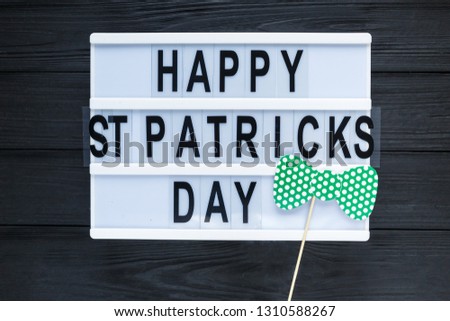 Lightbox with title Happy st Patrick Day and photobooth bow tie beard on wooden sticks on black wooden background. Creative background to St. Patricks Day Royalty-Free Stock Photo #1310588267