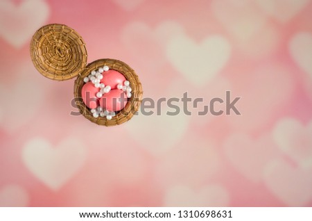 Tiny woven basket filled with pink egg shaped candies and pearly white dragee sprinkles on pink and white blurred heart background