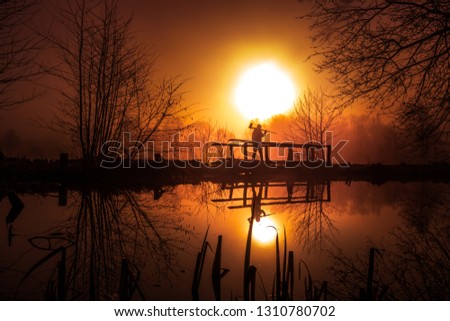 Photographer in an unreal landscape. he stands on an old wooden bridge tripod and camera shouldered and everything is reflected in the lake. the morning sun breaks orange in the thick fog