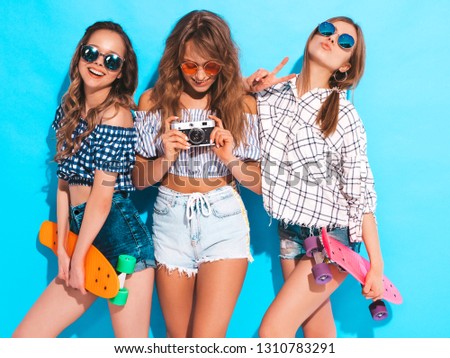 Three beautiful stylish smiling girls with penny skateboards in sunglasses.Women in summer hipster checkered shirt clothes near blue wall. Taking pictures on retro camera