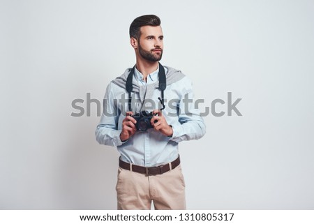 Confident photographer. Handsome young man in casual clothes is holding camera and looking away while standing on a grey background. Close-up. Hobby concept.