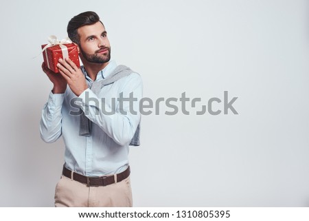 What is inside? Attractive stylish man is holding a gift box near his ear and trying to guess what is inside while standing on a grey background. Close-up image