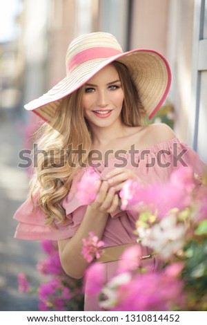 Beautiful young woman is laughing during her walk outside in the city. She is wearing a hat and pink summer dress, standing next to the pink flowers.