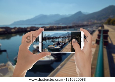 A man is making a photo of a small dock in Italy for boats and yachts with mountains on the background on a mobile phone
