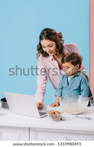 smiling mother using desktop while cooking together with little son on bicolor background
