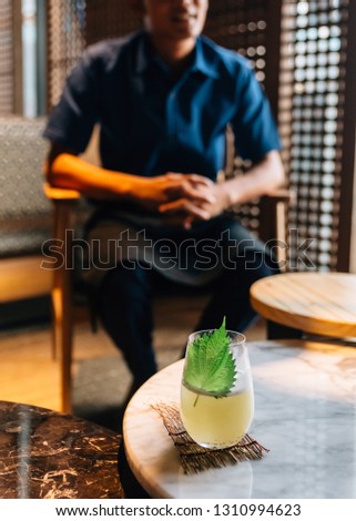 Japanese Yuzu Cocktail with Shiso Leaves (Green Perilla) in glass on marble low table with blur mixologist in background.