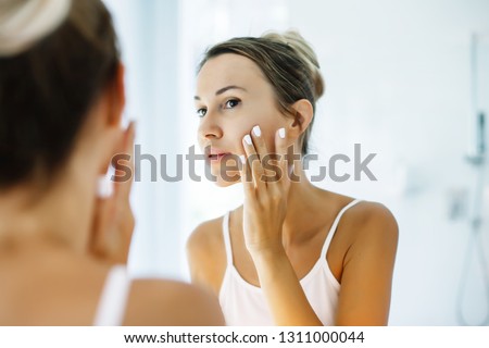 Photo of woman looking at mirror and touching her skin. Making everyday morning treatment in bathroom.