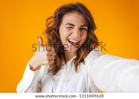 Portrait of a cheerful young woman wearing white shirt standing isolated over yellow background, taking a selfie