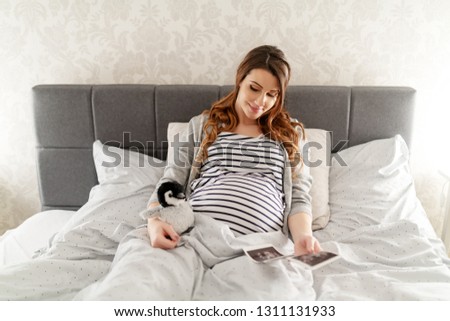 Happy cute brunette lying in the bed, holding penguin toy and looking at ultrasound picture of her baby.
