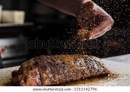 Raw piece of meat, beef ribs. The hand of a male chef puts salt and spices on a dark background, close-up. Royalty-Free Stock Photo #1311142796