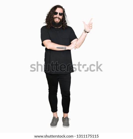 Young hipster man with long hair and beard wearing sunglasses with a big smile on face, pointing with hand and finger to the side looking at the camera.