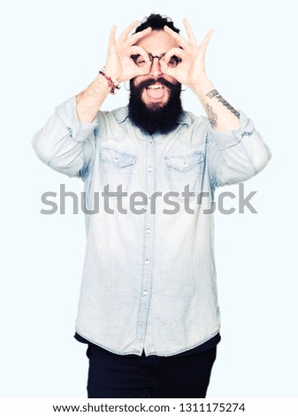 Young hipster man with long hair and beard wearing glasses doing ok gesture like binoculars sticking tongue out, eyes looking through fingers. Crazy expression.