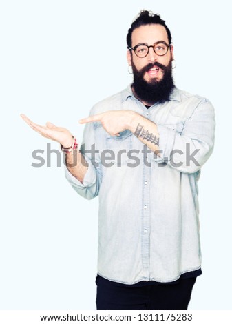 Young hipster man with long hair and beard wearing glasses amazed and smiling to the camera while presenting with hand and pointing with finger.