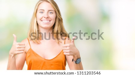 Beautiful young woman wearing orange shirt over isolated background success sign doing positive gesture with hand, thumbs up smiling and happy. Looking at the camera with cheerful expression, winner