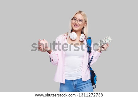Cost of college education. Portrait of a happy  female student  with piggy bank and cash money in  hands. The girl saves money for training in high school .Neutral grey background. Space for text Royalty-Free Stock Photo #1311272738