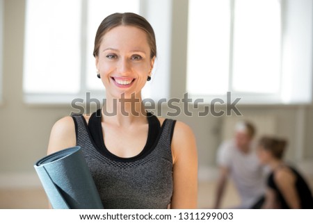 Portrait of excited female yoga teacher posing with fitness mat looking at camera, happy toned fitness instructor or coach in sportswear smiling making picture during training in yoga studio