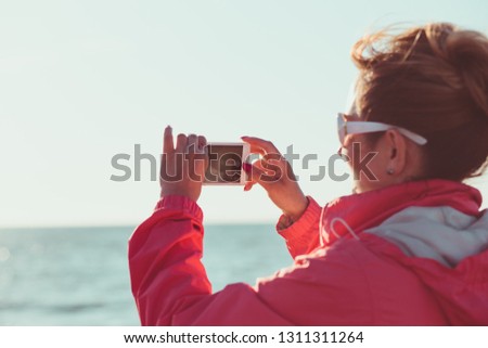 Young woman taking a photos using phone, looking at screen, standing outdoors, she is backlighted by sunlight with plain sky and sea in the background