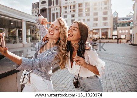 Wonderful blonde woman with long hairstyle making selfie with sister. Outdoor shot of attractive female friends posing with pleasure on city background.