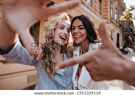 Laughing blithesome woman with dark hair walking down the street with friend. Carefree blonde girl posing on city background. Royalty-Free Stock Photo #1311329114