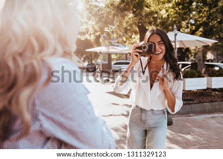 Happy girls making outdoor photoshoot. Brunette female photographer with camera taking pictures of friend.