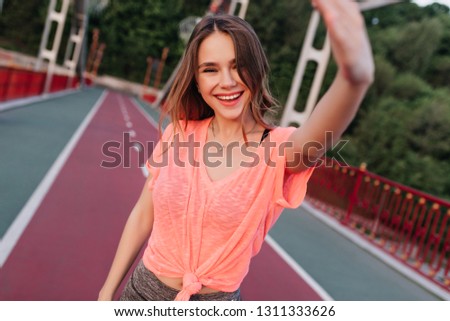 Excited brunette girl expressing positive emotions during photoshoot at stadium. Glad young lady making selfie at cinder path.