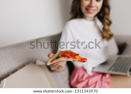 Charming woman using laptop and eating pizza with cheese. Indoor shot of relaxed girl in white t-shirt working with computer and enjoying fast food.