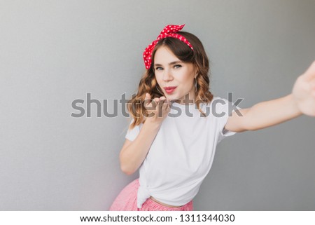 Glad caucasian lady sending air kiss on gray background. Good-humoured female model in red ribbon making selfie.