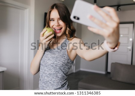Fashionable girl taking picture if herself while eating apple. Indoor shot of fascinating lady using phone for selfie.