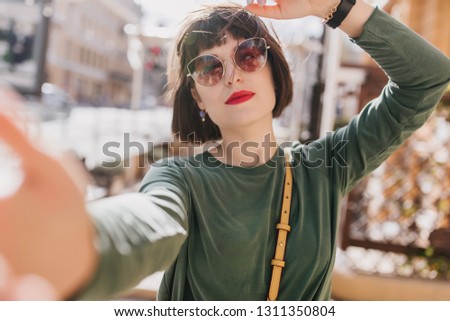 Fascinating young woman with red lips making selfie on city background. Outdoor shot of enthusiastic dark-haired girl wears sunglasses.