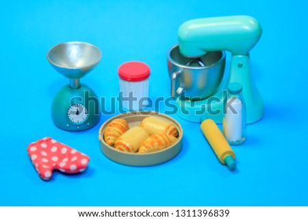 toy plastic cooking machine to prepare dough, bottle of milk, rolling pin, scales, oven glove and finished cakes on blue pastel minimal background