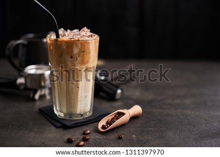 Ice coffee with cream in a tall glass and coffee beans, portafilter, tamper and milk jug on dark concrete table over black wooden background. Cold summer drink. Copy space for text. Selective focus. Royalty-Free Stock Photo #1311397970