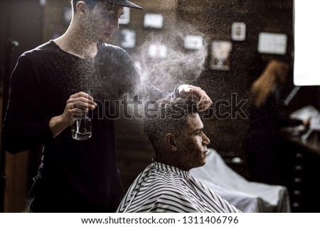 The barber dressed in a black clothes is doing the hair styling to the stylish black-haired  man sitting in the armchair in a barbershop Royalty-Free Stock Photo #1311406796