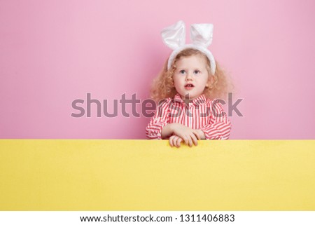 Funny little curly  girl in a striped red and white dress and bunny ears on her head stands  behind the yellow board against a pink wall