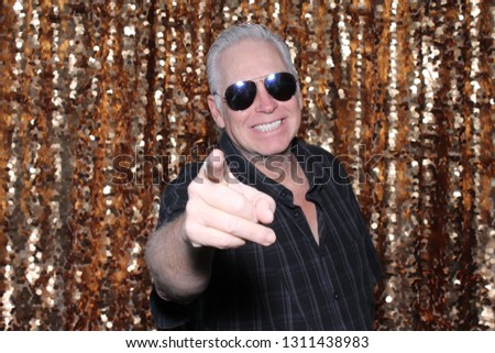 Man in a Photo Booth. A Caucasian man smiles and has fun posing in a Photo Booth with a Gold Sequin curtain. White male in sunglasses points to the camera and smiles.
 