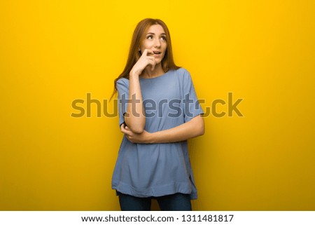 Young redhead girl over yellow wall background having doubts while looking up
