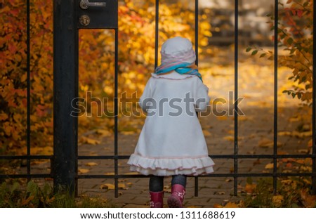 Little girl in white coat stands by the iron fence and looks at the garden in autumn