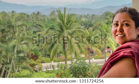Partial portrait of smiling middle-aged Mexican woman standing on balcony against green palm trees in blurred background, looking at camera, hair tied back, enjoying vacation in tropical day weather