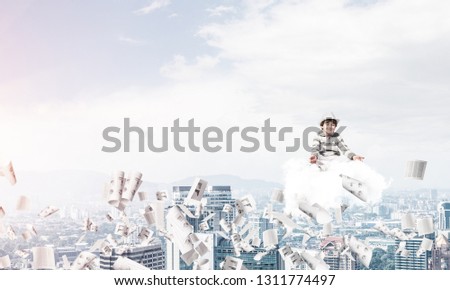 Young little boy keeping eyes closed and looking concentrated while meditating on cloud among flying papers with cityscape view on background.