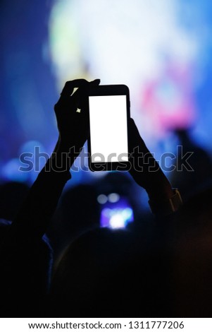Silhouette of music fan taking pictures,filming vertical video with smartphone on festival in night club.White empty mobile phone screen to place logo.Concert people partying in the club