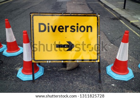 Yellow road sign "diversion" with a black arrow, stands on a blocked street on a gray asphalt road with traffic cones