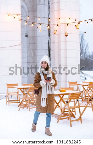 Photo of a pretty young woman in hat and scarf walking outdoors in winter snow drinking coffee using mobile phone.