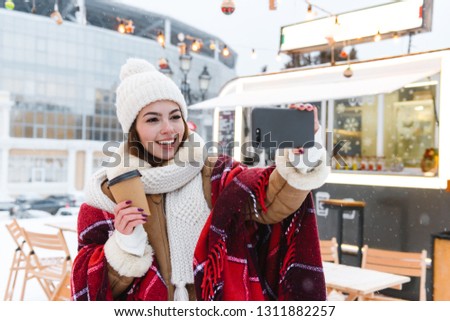 Photo of a pretty young woman in hat and scarf walking outdoors in winter snow drinking coffee take a selfie by mobile phone.