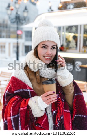 Photo of a pretty young woman in hat and scarf walking outdoors in winter snow drinking coffee talking by mobile phone.