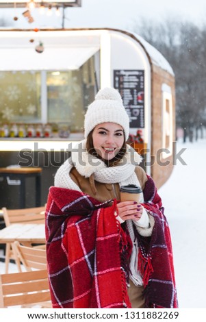 Photo of a pretty young woman in hat and scarf walking outdoors in winter snow drinking coffee.