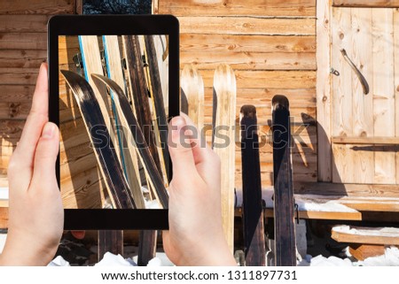 travel concept - tourist photographs of wide hunting skis near wall of wooden cottage in winter on smartphone in Smolensk region of Russia
