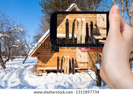 travel concept - tourist photographs of wide forest skis in front of wooden cottage in sunny winter day on smartphone in Smolensk region of Russia